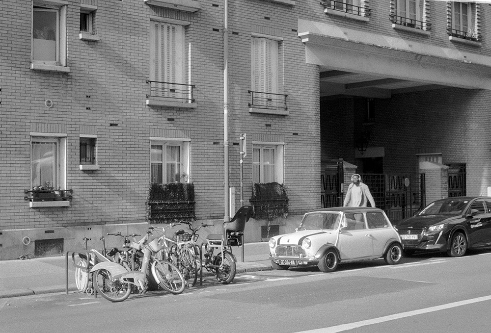 horizontal black and white photo of brick building across the street with an old classic Mini Cooper parked in front