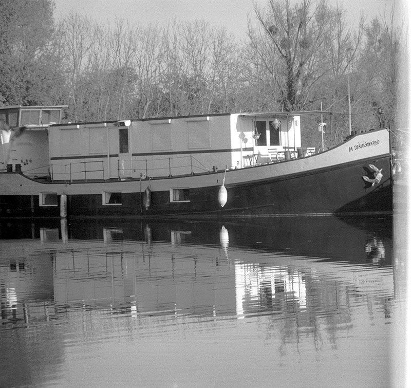 square-ish black and white photo of a boat on a river, the right of the image is cropped due to a light leak